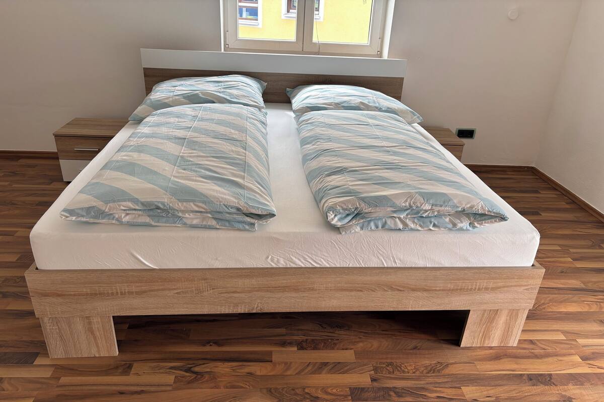 A double bed with a wooden frame and a mattress, two pillows, a nightstand, and a window with a view of buildings.