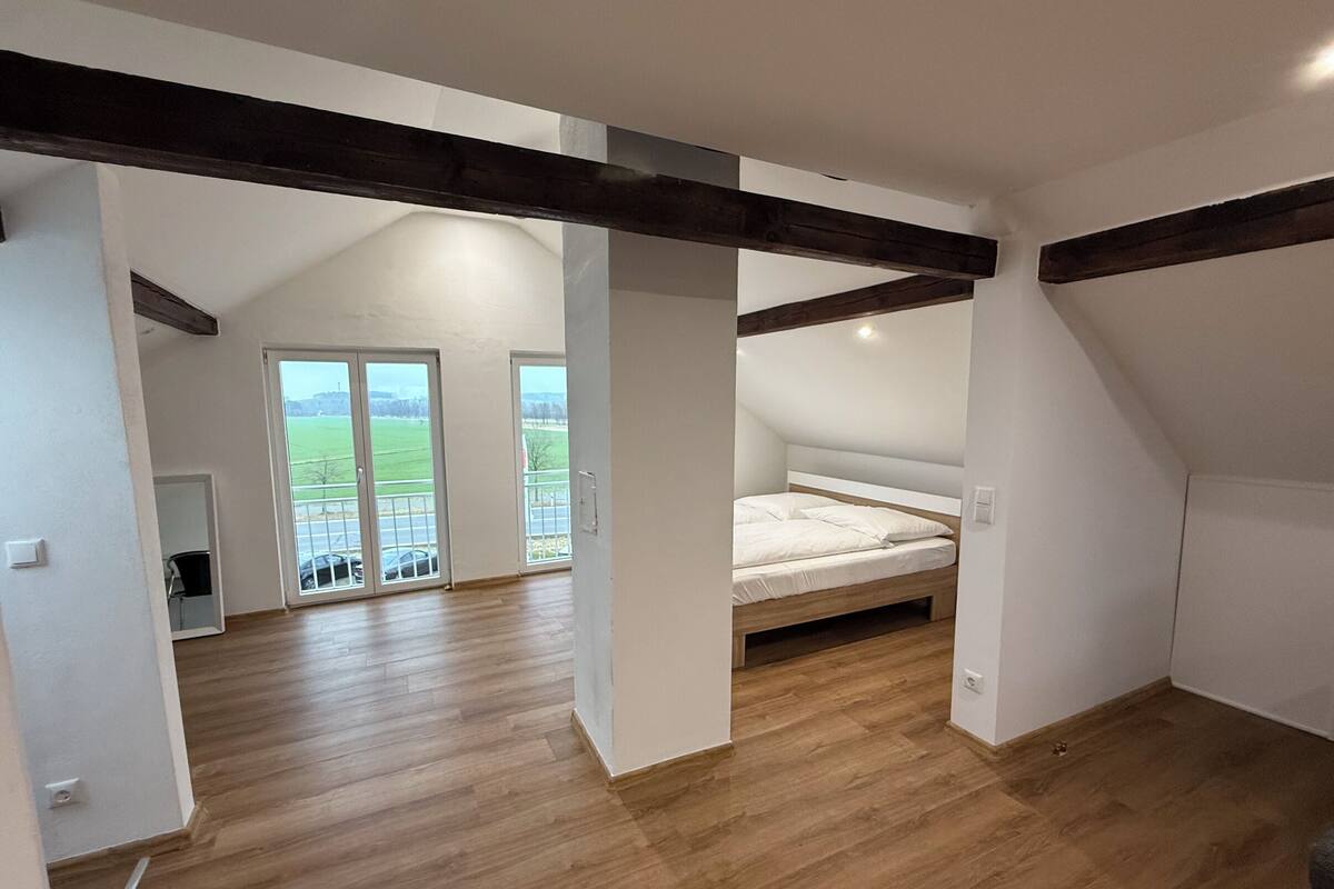 A room with a sloped ceiling, wooden beams, a bed, and a balcony with a view of a green landscape.