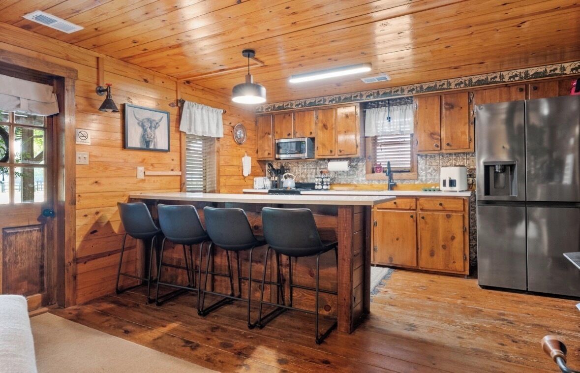 A wooden cabin interior with a kitchen area featuring a refrigerator, microwave, and bar stools.