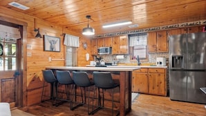 A wooden cabin interior with a kitchen area featuring a refrigerator, microwave, and bar stools.