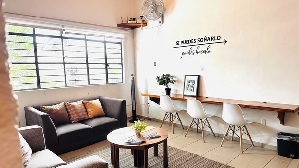 A modern living room with a sofa, coffee table, and chairs. There is a window with blinds, a wall-mounted fan, and a shelf with a plant and picture frame. - Espacio Libertad  (Guadalajara)