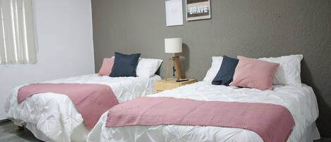 Habitación de hotel con dos camas, cada una con un edredón blanco y mantas rosas. Hay una mesita de noche con una lámpara y un cuadro en la pared.