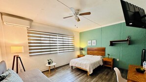 A hotel room with a bed, a desk, a chair, a ceiling fan, a TV, a window with blinds, a lamp, and a small table with a plant. - Espacio Libertad  (Guadalajara)