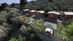 A row of wooden cabins with metal roofs, surrounded by a grassy area and trees, set against a hilly backdrop. - İda Tahtalıköy Bungalov (Edremit)