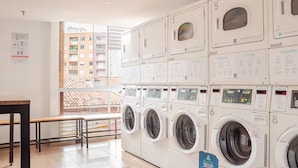 Une laverie équipée de plusieurs machines à laver et de plusieurs séchoirs, une affiche indiquant les heures d’ouverture, et une vue sur un immeuble d’habitation à travers une porte vitrée.