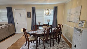 Un comedor con mesa de madera, sillas y alfombra floral. Se observa una araña colgante sobre la mesa y un cuadro en la pared.