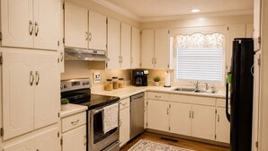 Una cocina con armarios blancos, estufa, lavavajillas, refrigerador y ventana con persianas.