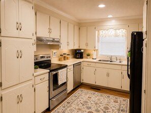 A kitchen with white cabinets, a stove, a dishwasher, a refrigerator, and a window with blinds. - Hot Tub + Fenced Yard | Family & Pet Friendly (Dothan)