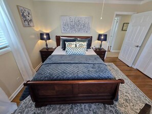A bedroom with a wooden bed, two bedside tables with lamps, a large floral artwork above the bed, and a door to the right. - Hot Tub + Fenced Yard | Family & Pet Friendly (Dothan)