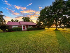 A single-story house with a red roof and a front porch, surrounded by a well-maintained lawn and a large tree. - Hot Tub + Fenced Yard | Family & Pet Friendly (Dothan)