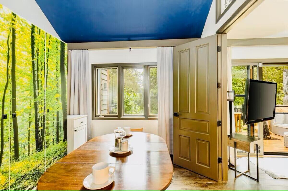 A room with a large window, a painting of a forest, a wooden table with cups, and a TV.