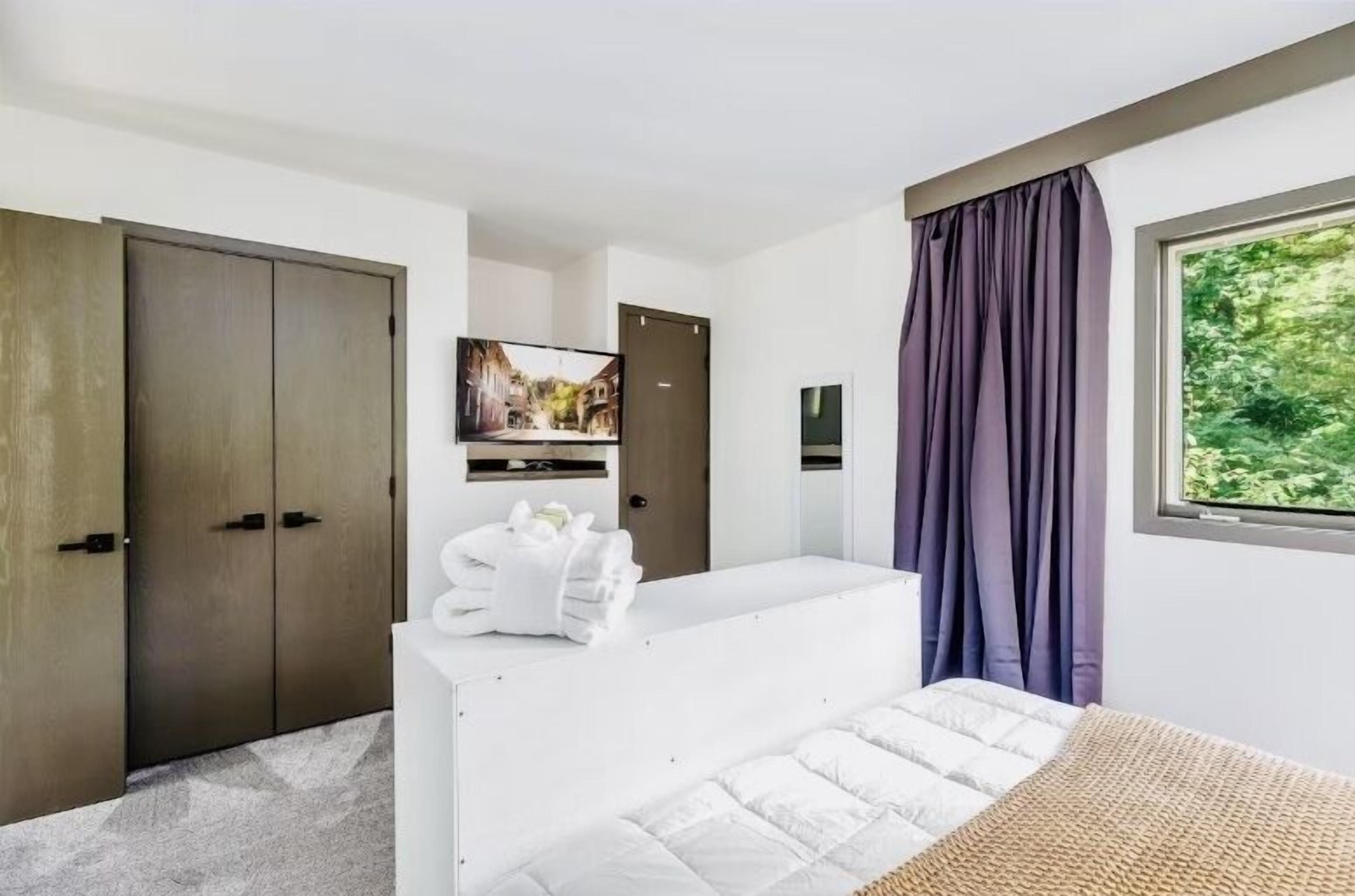 Une chambre d’hôtel moderne équipée d’un lit, d’un téléviseur à écran plat fixé au mur, d’une armoire et d’une fenêtre avec des rideaux.