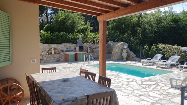 Eine Außenpatio mit Pool, Steinböden, einem Tisch mit Stühlen, einer Holzpergola und einer Steinmauer mit integriertem Grill.