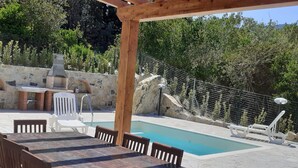 Ein Außenpoolbereich mit einer Holzpergola, einer Steinmauer und einer Grillgelegenheit.