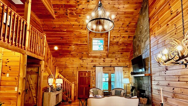 Interior de una cabaña de troncos con escalera de madera, candelabro, chimenea y un rincón de estar acogedor.