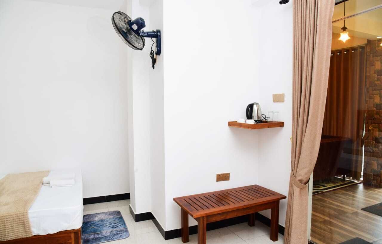 Una habitación pequeña con una cama, un banco, un ventilador de pared y un estante con una tetera.