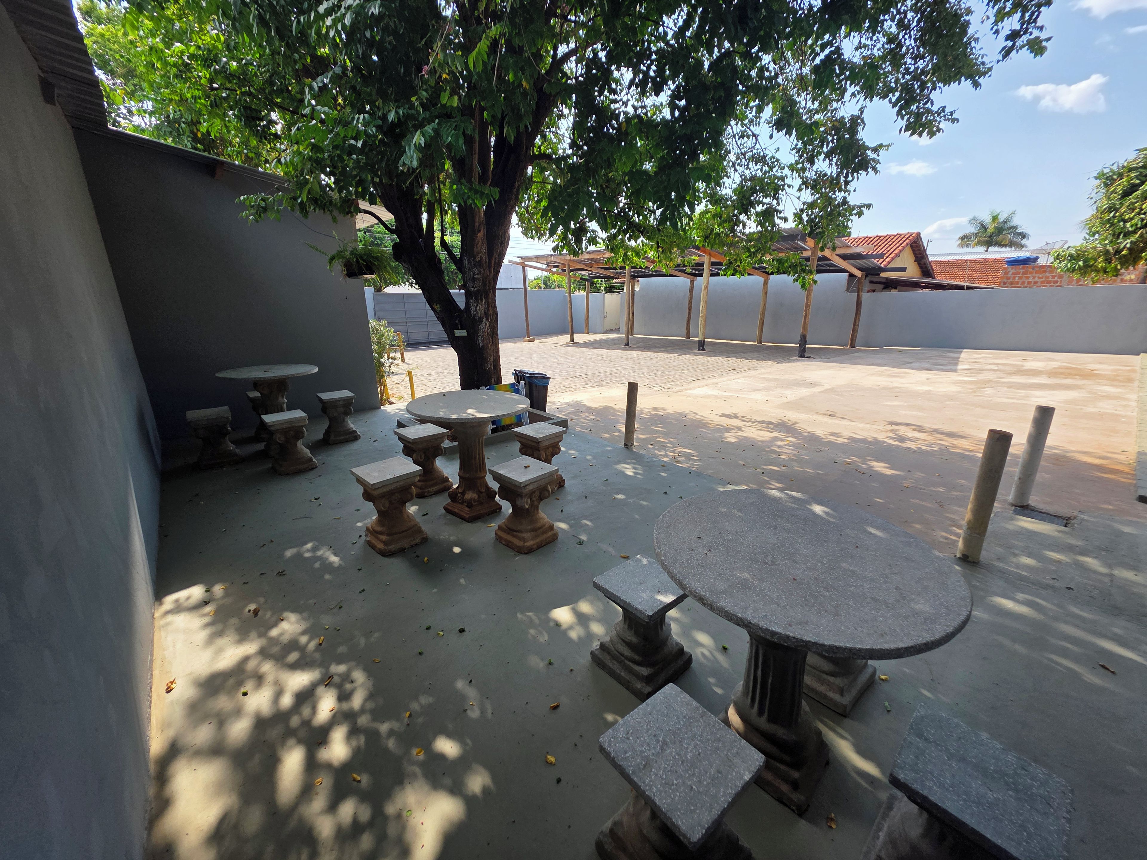 Een terras buiten met een ronde tafel en meerdere banken, omringd door een lage muur en bomen.