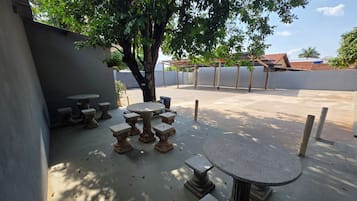 Een terras buiten met een ronde tafel en meerdere banken, omringd door een lage muur en bomen.