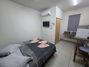 Un dormitorio con cama, televisor, aire acondicionado, puerta de madera, pequeña cocina y baño.