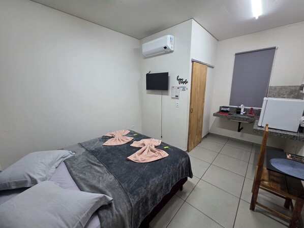 Un dormitorio con cama, televisor, aire acondicionado, puerta de madera, pequeña cocina y baño.