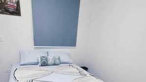 Een net opgemaakt bed met een bedsprei met dessin, een raam met zonwering en een klein schilderij aan de muur.