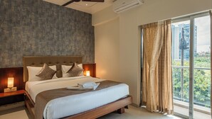 A modern bedroom with a large bed, wooden headboard, bedside lamps, and a view of greenery through a large window. - StayBird - Azure Suites Business Hotel (Pune)