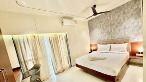 A hotel room with a large bed, a bedside table with a lamp, a chair, a ceiling fan, and a window with curtains. - StayBird - Azure Suites Business Hotel (Pune)
