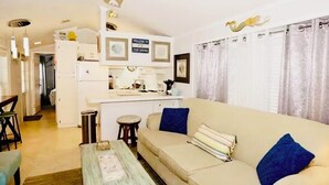 A living room with a sofa, a coffee table, and a ceiling fan. There is a kitchen area with a refrigerator and a sink.