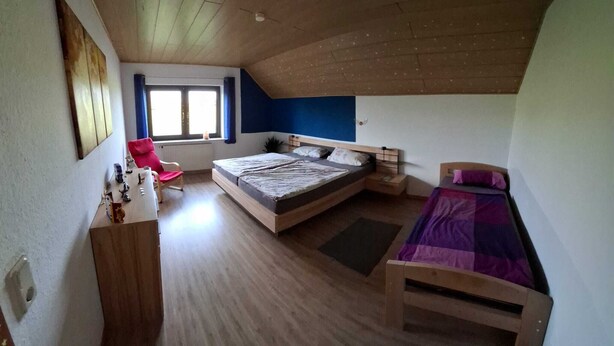 A bedroom with a sloped ceiling, a bed, a chair, and a desk with a lamp. - Large, cozy and family-friendly vacation apartment with balcony and garden (Bischbrunn)