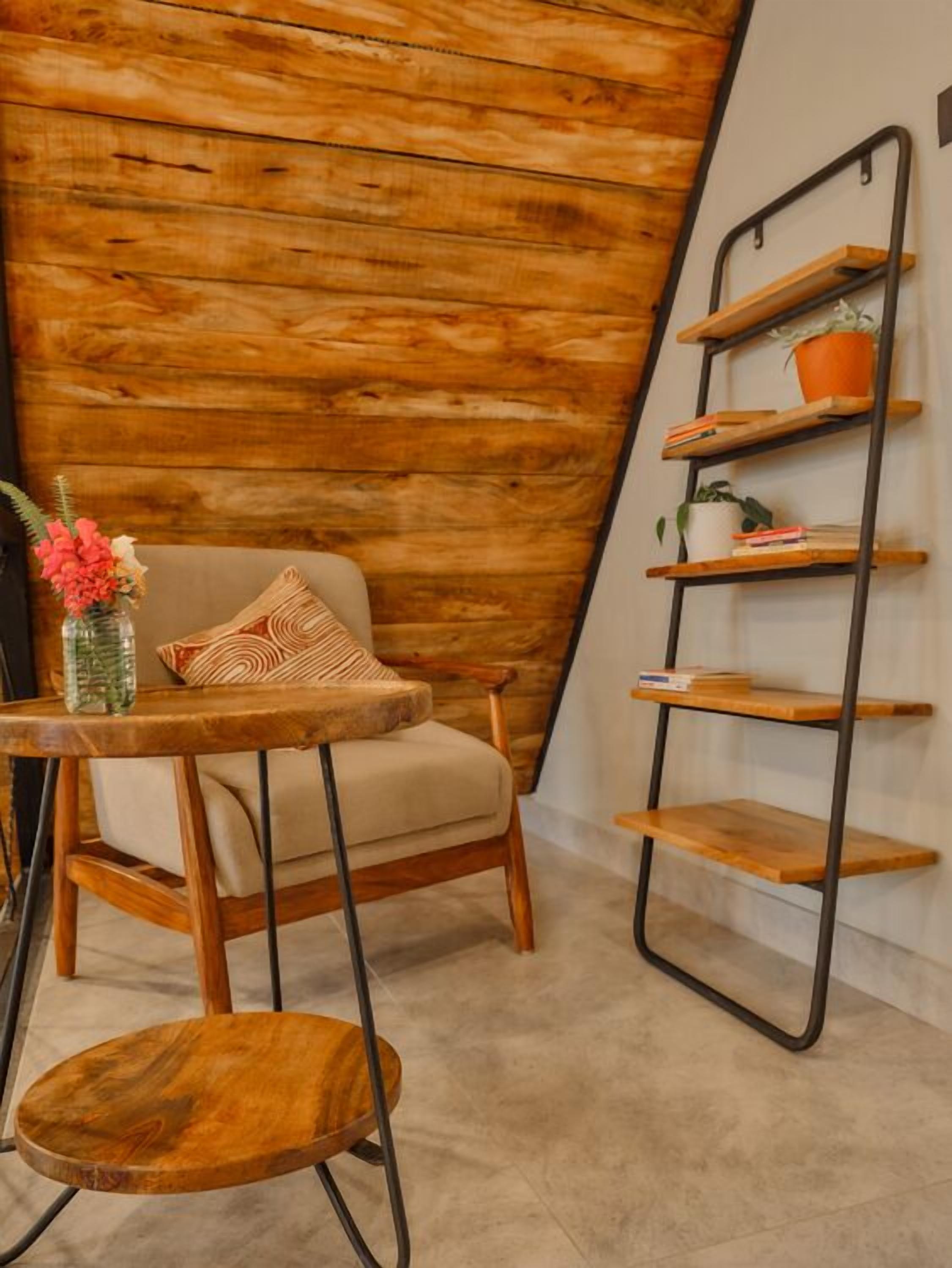Una habitación acogedora con techo de madera inclinado, una silla de madera con cojín, una mesita redonda de madera y un estante metálico con plantas en macetas.