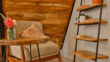 Una habitación acogedora con techo de madera inclinado, una silla de madera con cojín, una mesita redonda de madera y un estante metálico con plantas en macetas.