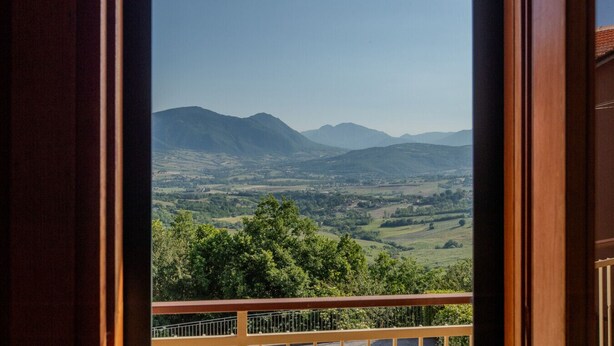 Blick von einem Balkon mit Holzgeländer auf ein Tal mit Bergen im Hintergrund.