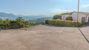 Ein gepflasterter Außenbereich mit einem Metallzaun, ein weißes Gebäude mit einem Ziegeldach und Blick auf ferne Berge.