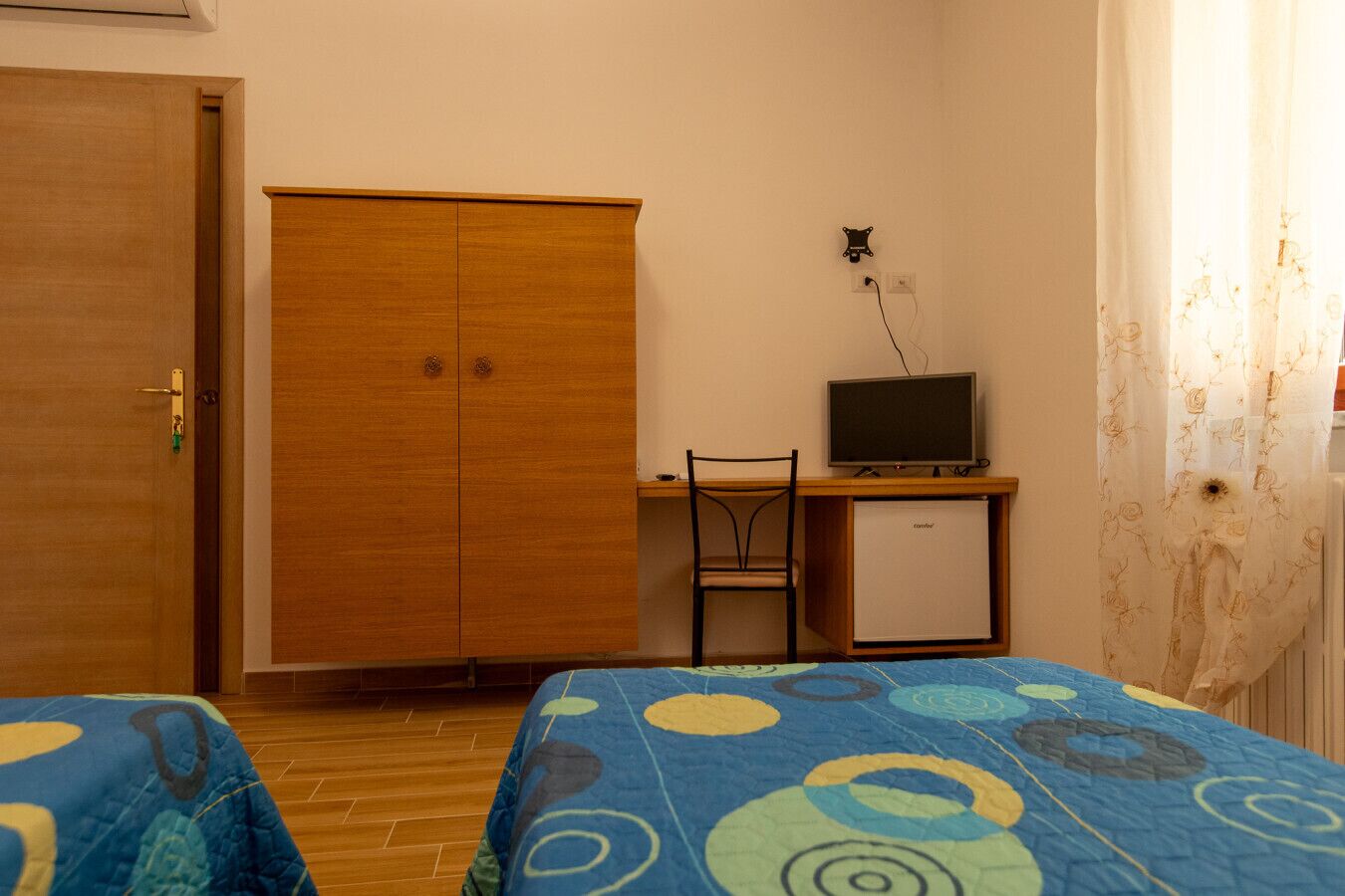 Een kamer met twee bedden, een houten garderobe, een bureau met een computer en een kleine koelkast.