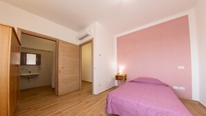 Une chambre avec un plancher en bois, un lit recouvert d’une couverture rose, une lampe de chevet et une armoire en bois.
