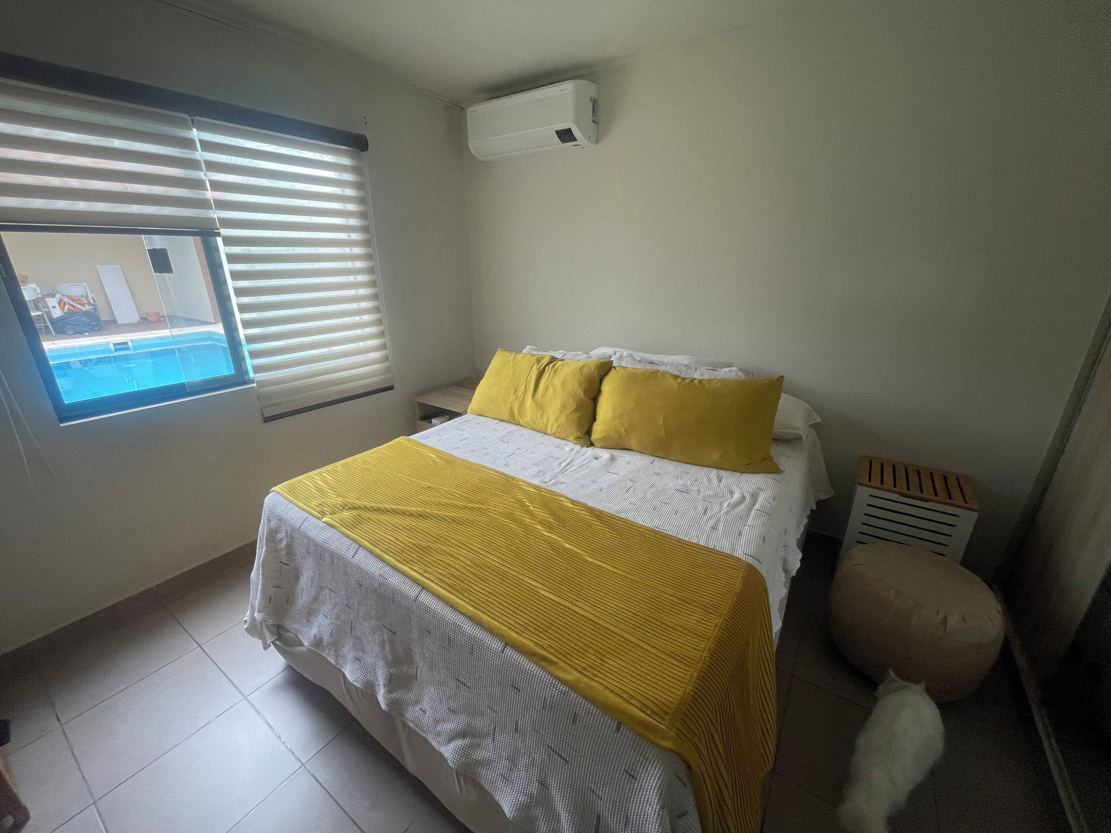 A bedroom with a bed, a window with blinds, an air conditioning unit, and a small heater.