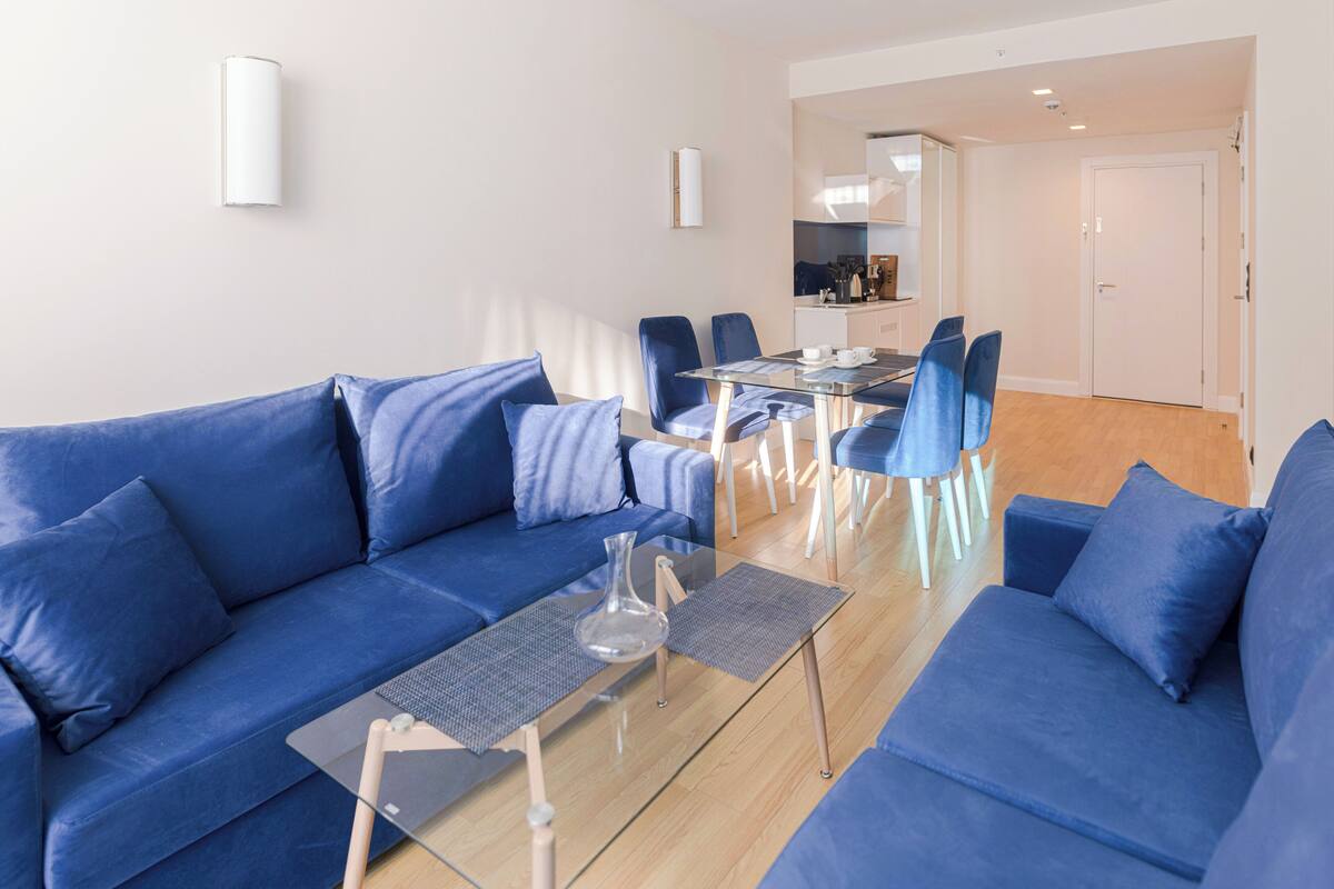 A modern living room with a blue sofa set, a glass coffee table, and a dining area with chairs and a table.