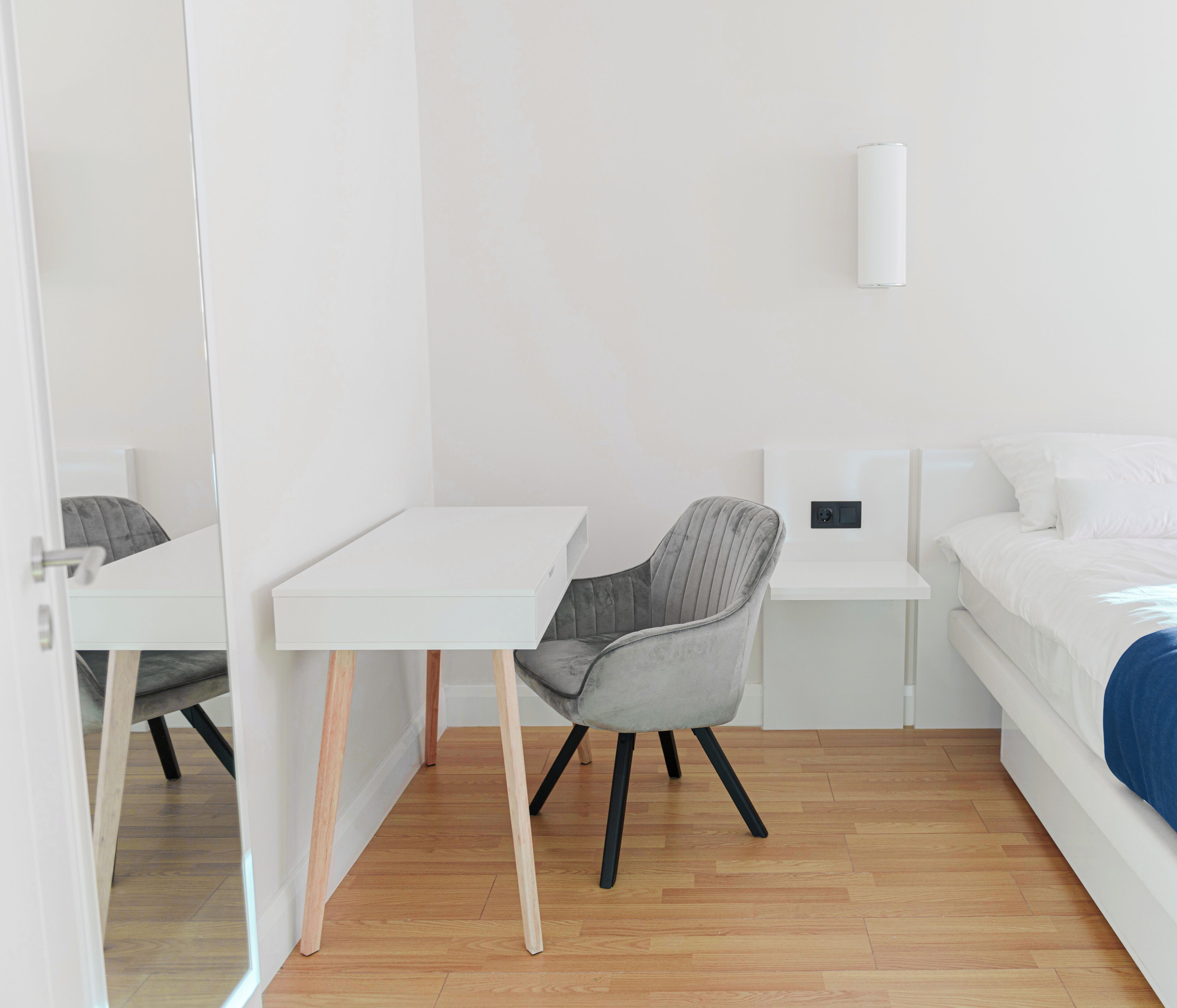A modern, minimalist room with a white desk, a grey chair, a white bedside table, and a bed with white bedding.