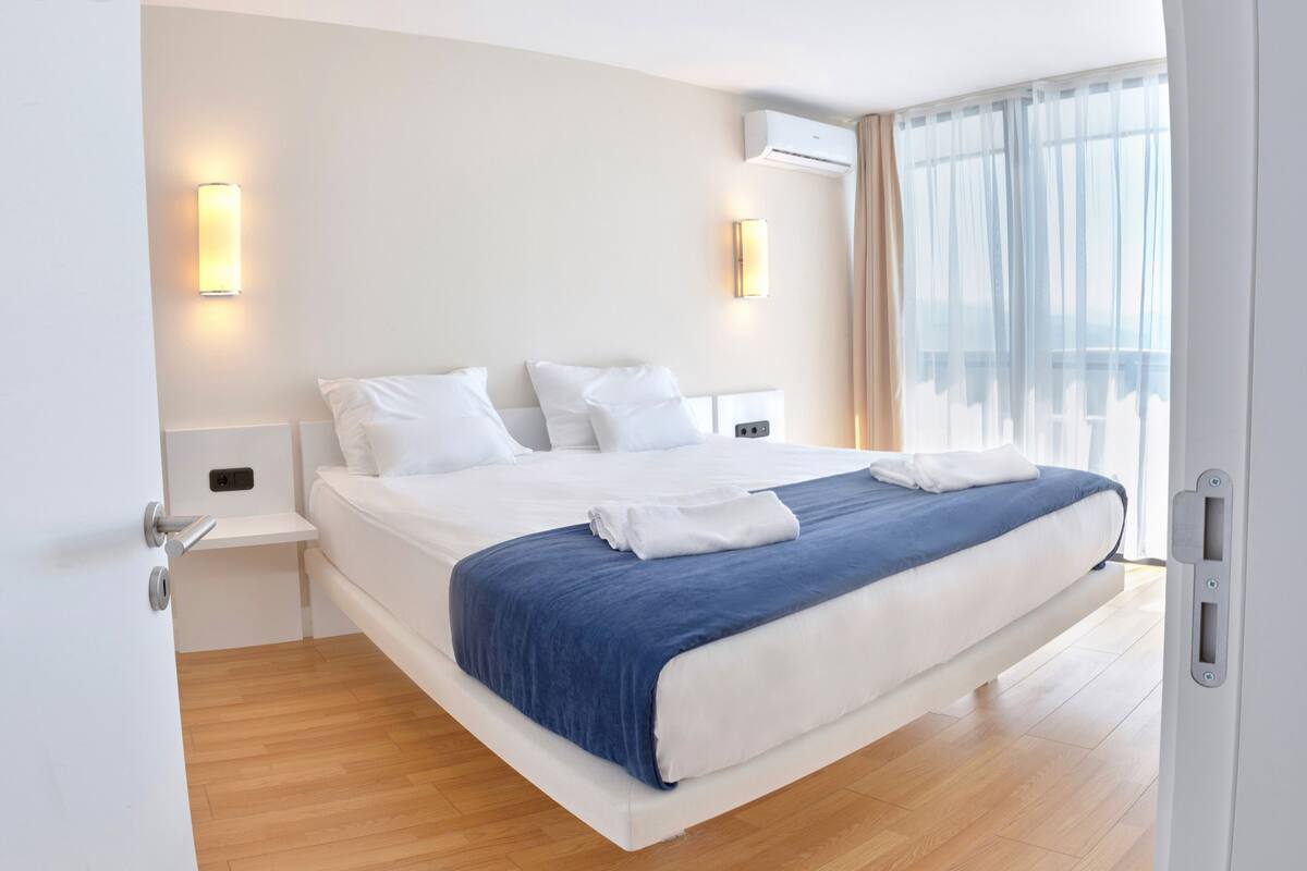 A modern hotel room with a large bed, white walls, and a wooden floor.