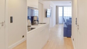 Una cocina moderna con estufa integrada, cafetera y pileta. En la zona de estar contigua, hay un sofá azul y una ventana con cortinas.