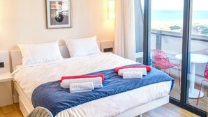 Habitación de hotel con una cama grande, vista a la playa y un balcón con sillas rojas.