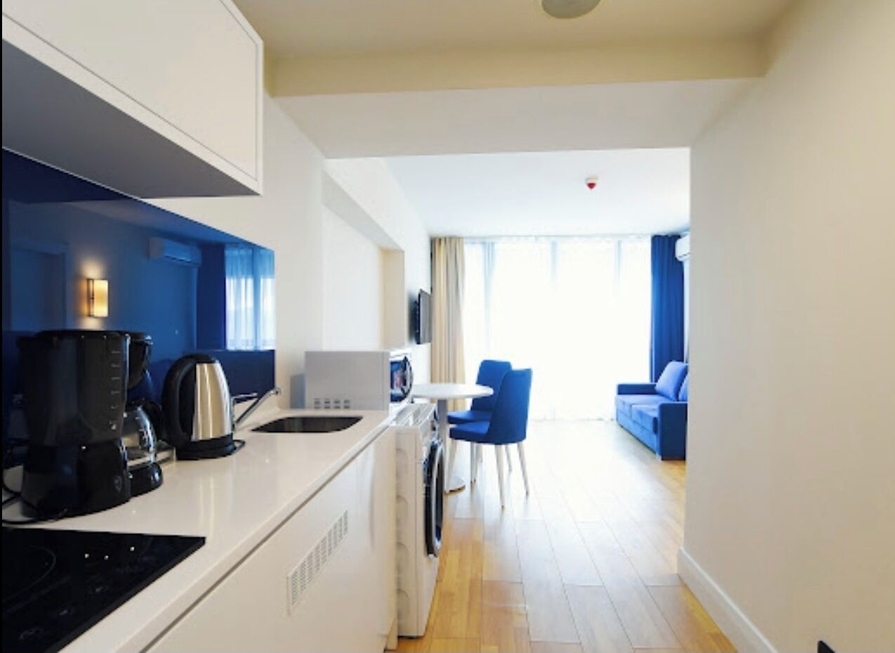 A modern kitchen with a coffee maker, kettle, and microwave. There is a dining area with a table and chairs. The room has a blue accent wall and a large window.