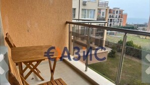 Un balcón con mesa y silla de madera, baranda de cristal y vistas a edificios y follaje.