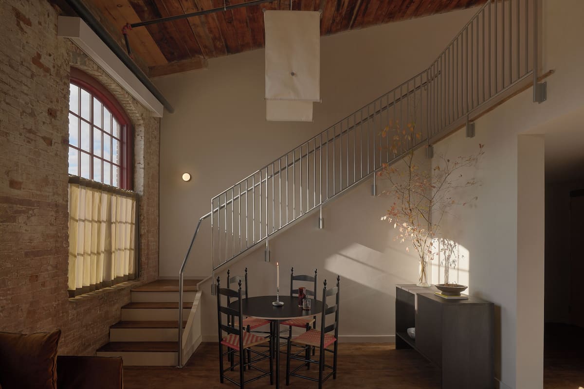 A room with a staircase, a dining table with chairs, a vase with branches, and a brick wall.