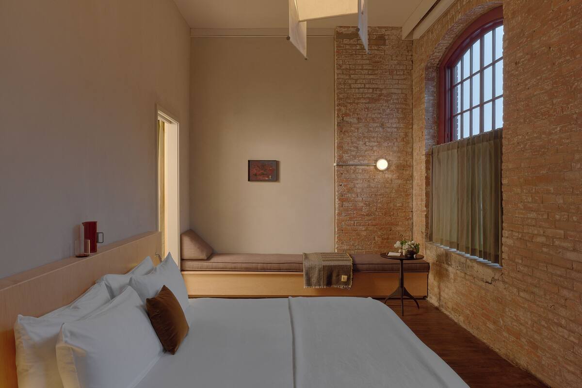 A hotel room with a bed, a small table with a lamp, a window with curtains, and a brick wall.