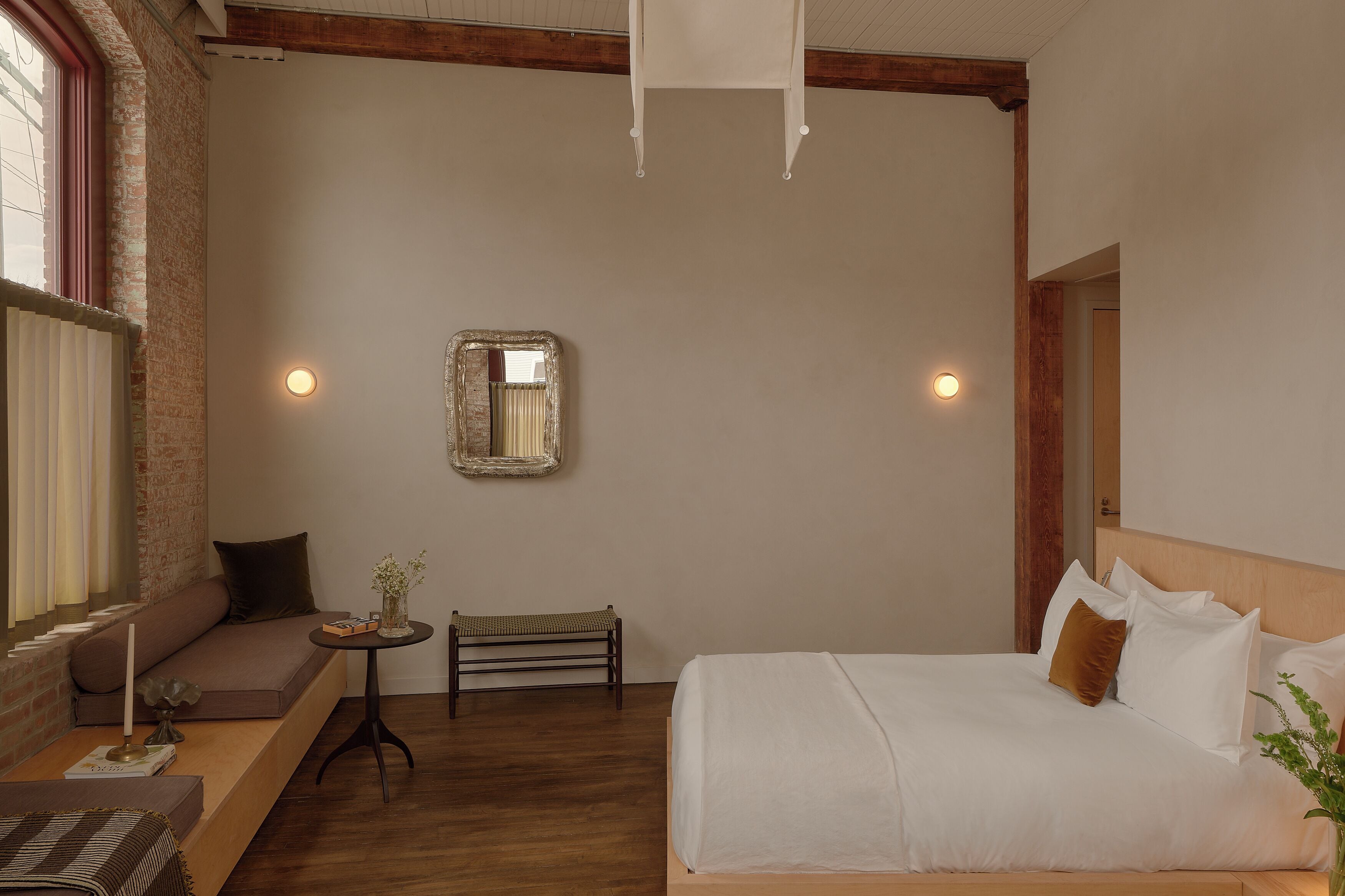 Quarto com cama, um banquinho, uma mesinha, um espelho e uma janela com persianas.