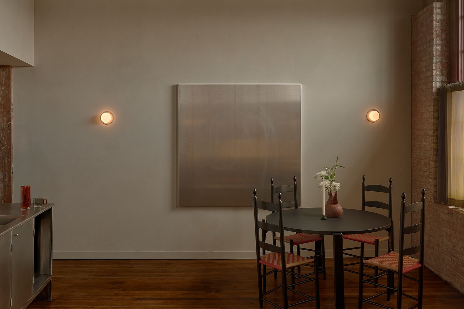 A dining area with a round table, chairs, a vase with flowers, and wall-mounted lights.