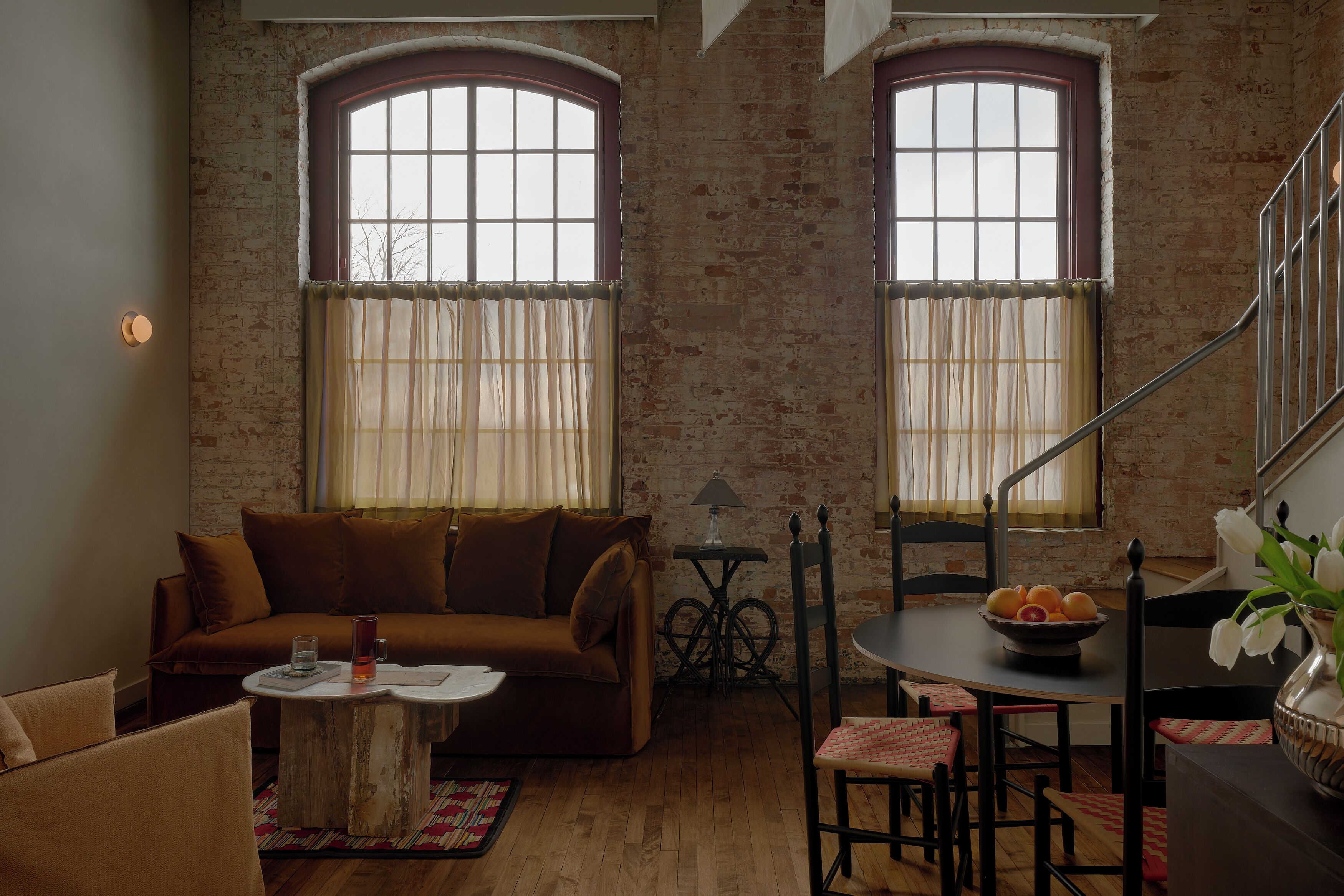 A living room with a brown sofa, a coffee table, a dining table with chairs, and two windows with curtains.