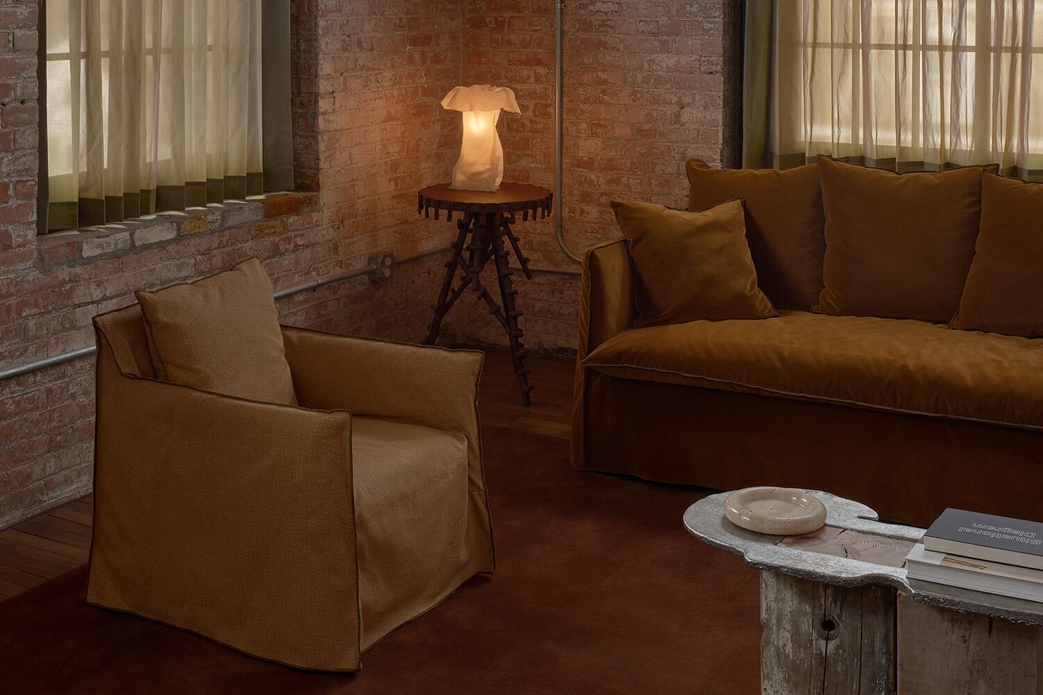 A room with a brick wall, a sofa, a chair, a small table with a lamp, and a window with curtains.