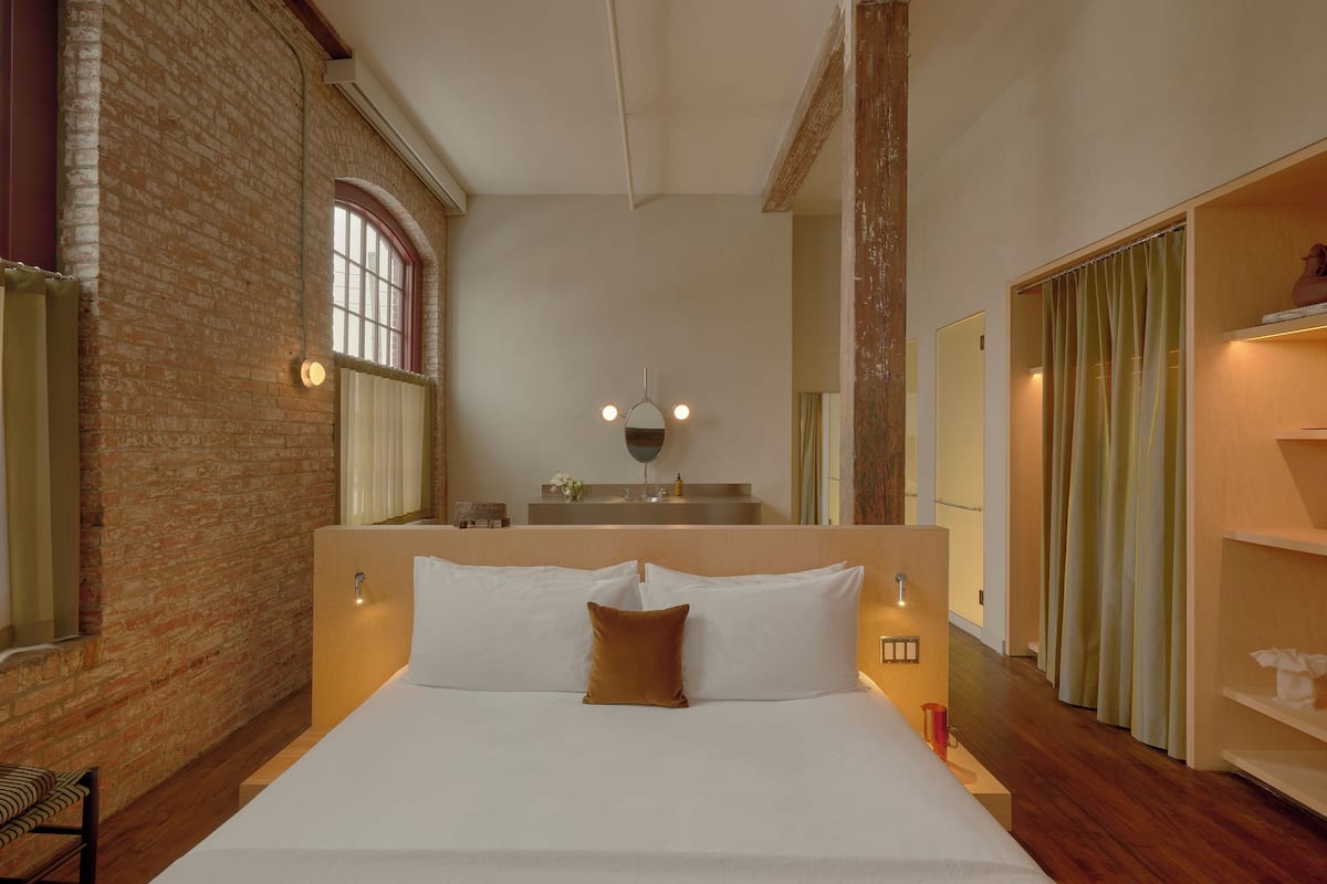A hotel room with a large bed, a brick wall, a mirror, a bathroom with a shower curtain, and a wooden floor.